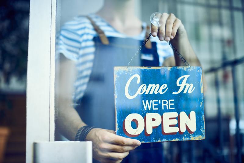 A figure in overalls holds a blue sign in a shop window that reads "Come in, we're open"