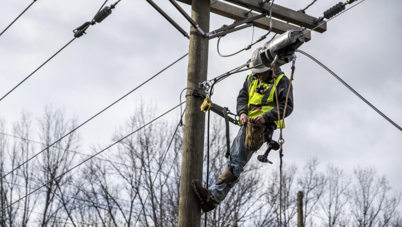 A line worker in a white hard hat and bright yellow visibility vest is securely attached to a power line, and is working on them